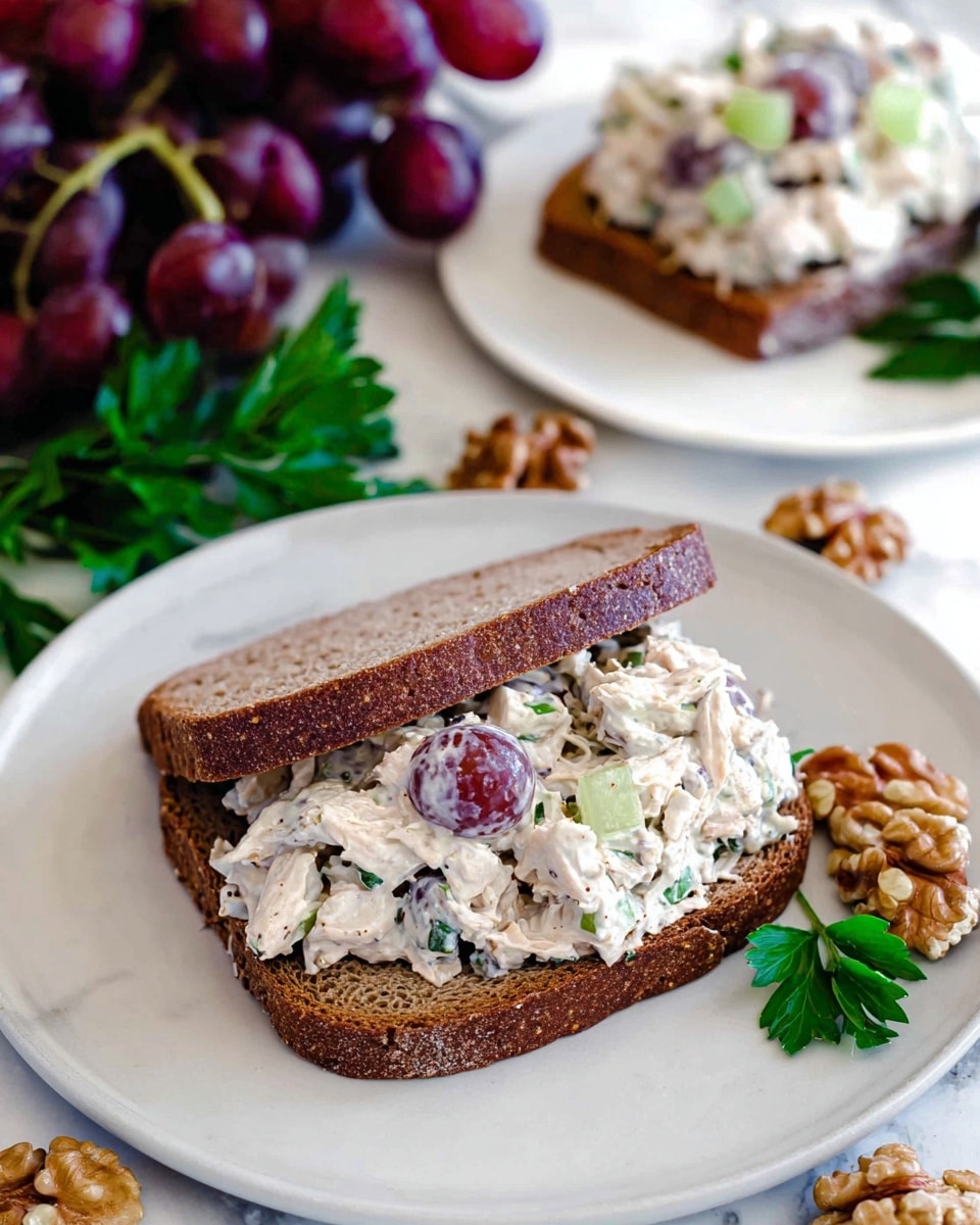 A sandwich with two slices of dark brown bread, the bottom slice topped thickly with a creamy chicken salad mixture containing visible chunks of shredded chicken, red grapes, green celery pieces, and herbs, all in a white dressing, sits on a white plate. In the background, another plate holds an open-faced sandwich with the same filling on dark brown bread. Around the plate are scattered walnut pieces, some red grapes on the vine, and a sprig of fresh green parsley, all placed on a white marbled surface. photo taken with an iphone --ar 4:5 --v 7