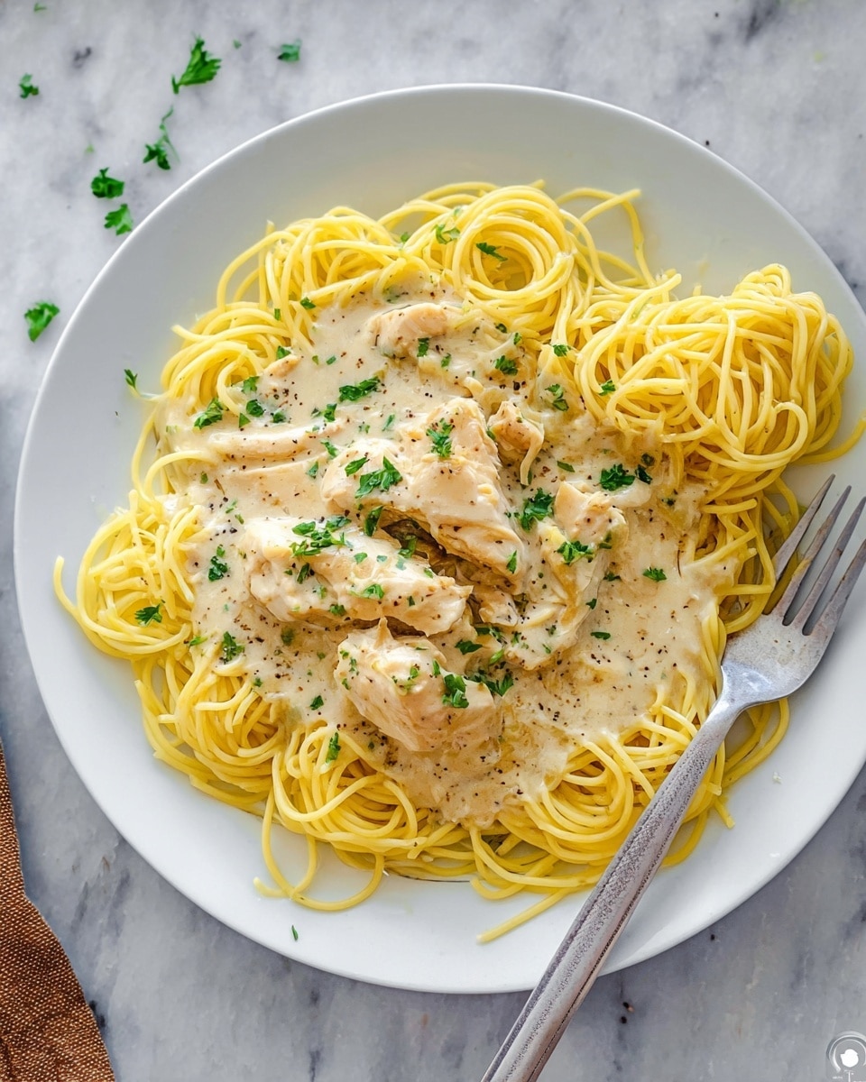 A white plate holds a serving of yellow spaghetti arranged in loose coils forming the bottom layer. On top, there are pieces of white chicken covered in a creamy, light beige sauce that looks smooth with small specks of black pepper visible. Bright green small parsley leaves are scattered on the sauce, adding color contrast. A silver fork rests on the right side of the plate, partially twirling some spaghetti. The whole scene is set against a white marbled texture. photo taken with an iphone --ar 4:5 --v 7