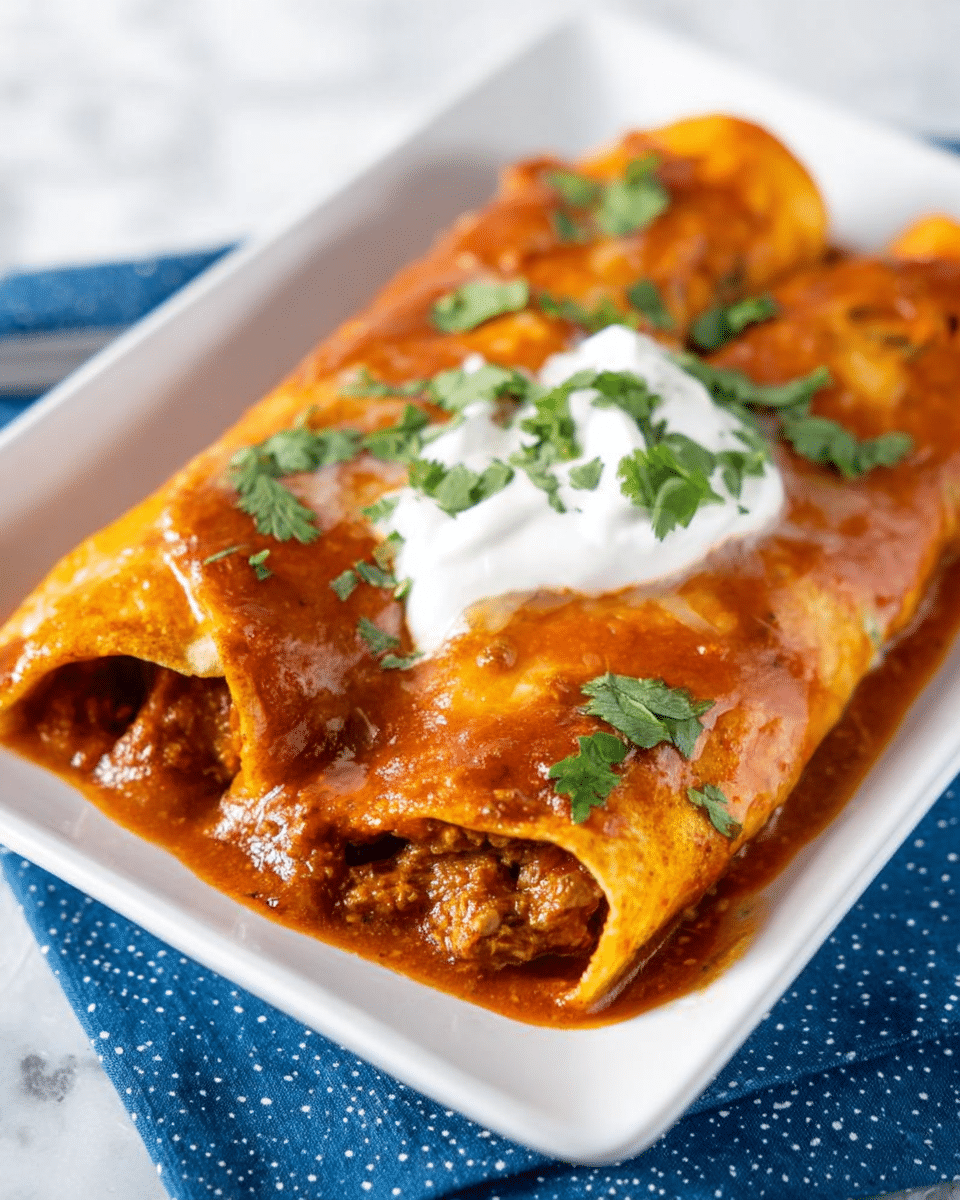 The image shows two rolled enchiladas placed closely on a white rectangular plate. The enchiladas are covered with a smooth, rich red sauce that has a slightly oily shine, with bits of ground meat visible through the sauce near the edges. On top of the middle enchilada, there is a dollop of white sour cream with a few green cilantro leaves placed decoratively on it and scattered around the plate. The plate sits on a blue cloth with small white dots, all set on a white marbled surface in the background. Photo taken with an iphone --ar 4:5 --v 7