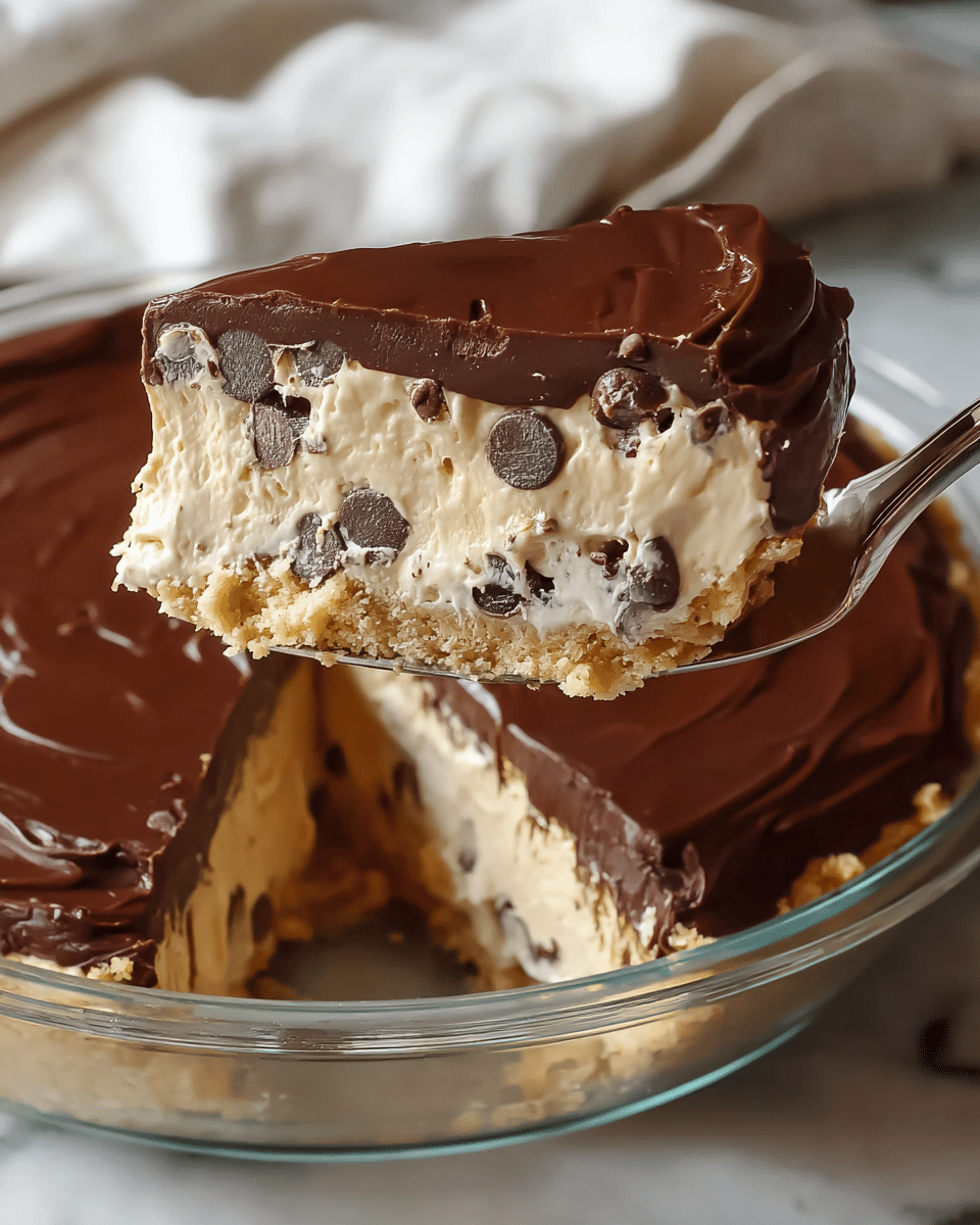 The image shows a close-up of a slice of a layered dessert held by a spoon above a white marbled surface. The dessert has three layers: the bottom layer is a crumbly, light tan crust; the middle layer is thick, creamy, pale beige filling with chocolate chips scattered throughout; the top layer is a smooth, glossy dark chocolate ganache spread evenly. The dessert is in a clear glass pie dish with more slices visible inside, and a white cloth is partially seen in the background. photo taken with an iphone --ar 4:5 --v 7