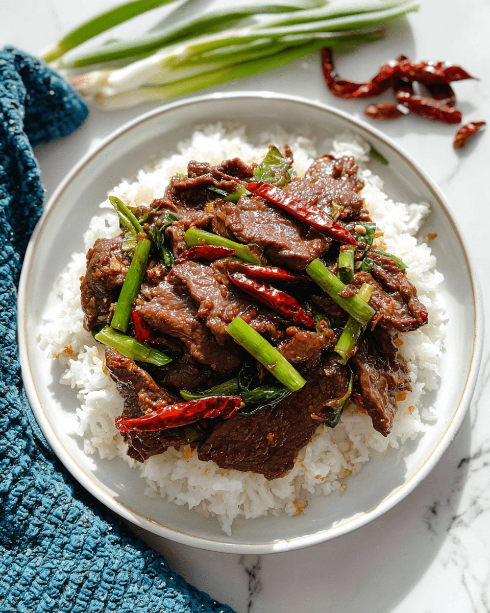 The image shows a white plate filled with a bed of white rice as the bottom layer, topped with stir-fried beef strips that are brown and glossy with a rich sauce. Mixed in with the beef are bright green pieces of sliced green onions and whole dried red chili peppers, adding a pop of color and texture contrast. The dish has a shiny, slightly oily look from the sauce, and the ingredients are arranged centrally on the plate. The plate is placed on a white marbled surface, with some green onions and dried red chili peppers visible in the background, and a blue textured cloth on the side. photo taken with an iphone --ar 4:5 --v 7