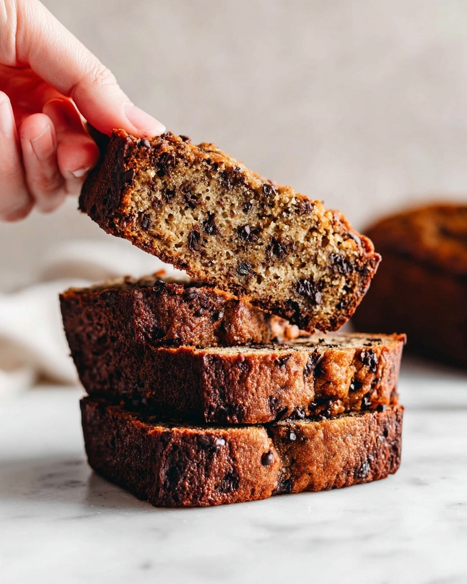 A single slice of moist banana bread is shown on a white marbled surface, with a golden brown crust that is slightly darker on the edges. The inside of the bread is light brown and textured, dotted generously with melted dark chocolate chips throughout. The top layer shows a slightly rough, crumbly texture with visible chocolate bits. Another slice is partially visible at the bottom right corner, and a blurred cooling rack is in the background. photo taken with an iphone --ar 4:5 --v 7