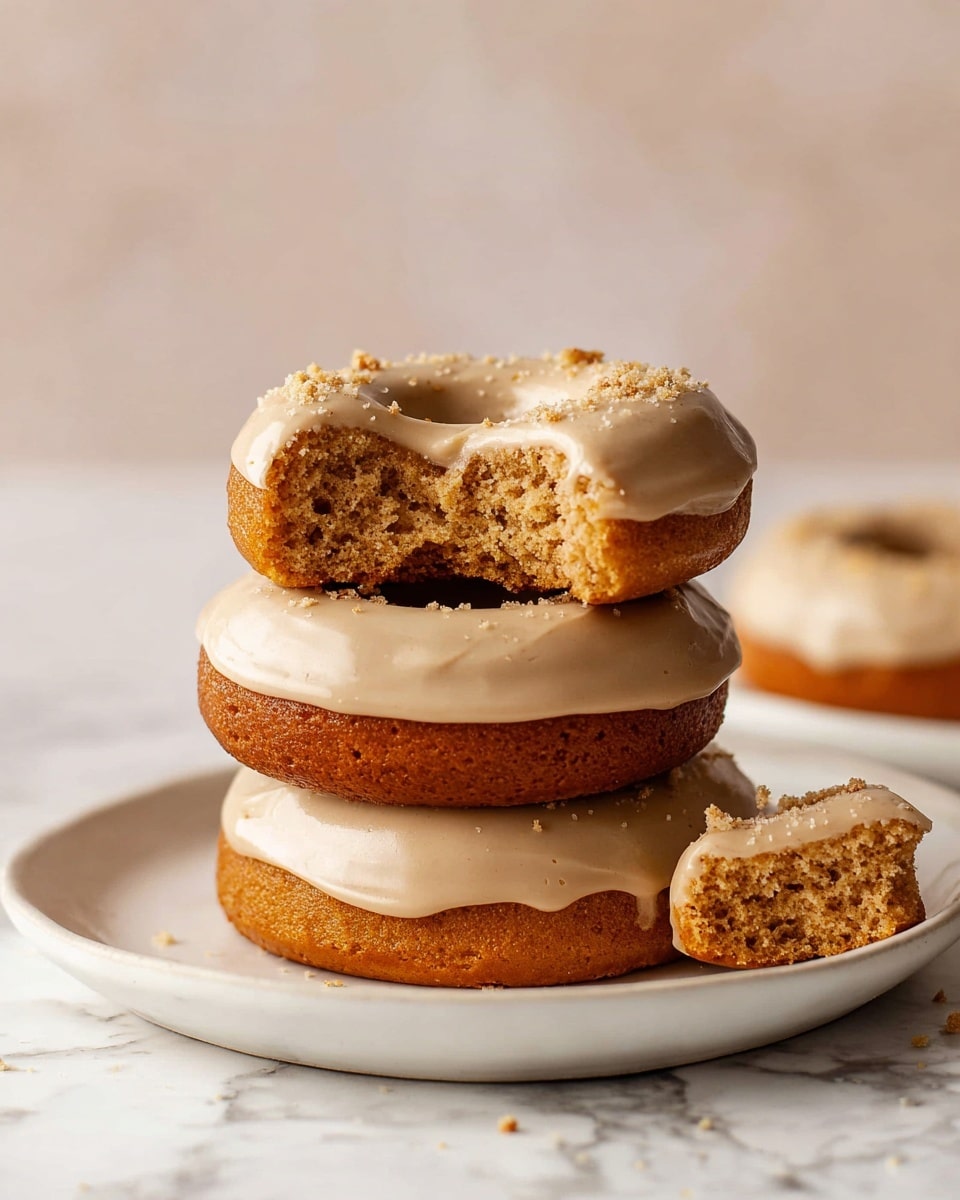 The image shows a stack of three thick, round donuts on a white plate with a white marbled background. Each donut has a golden-brown base with a smooth, creamy tan frosting layer that covers the top. The second donut from the top has small crumbs sprinkled on the frosting. The top donut has a bite taken out of it, revealing a soft, moist internal texture of a rich golden-brown color. A small piece of this bitten donut lies in front of the plate. The whole scene is softly lit, highlighting the texture and color contrast between the golden donut and its light frosting. photo taken with an iphone --ar 4:5 --v 7