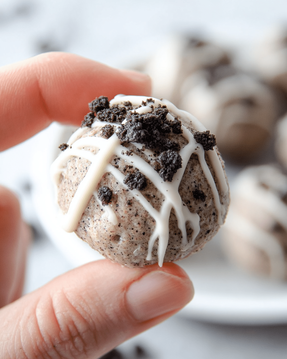 A close-up of a small round dessert ball held between a woman's thumb and forefinger, showing a light grayish-brown base with a slightly rough texture mixed with tiny dark specks. On top are thin white icing drizzle lines crossing each other in a grid pattern. Scattered over the icing are small chunks of dark brown cookie crumbs adding a crumbly texture. The background shows a soft-focus white marbled texture and a white plate with similar dessert balls. photo taken with an iphone --ar 4:5 --v 7