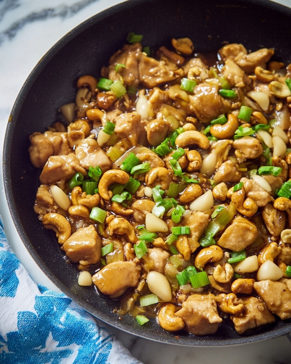 The image shows a close-up of a black pan filled with cooked bite-sized pieces of light brown chicken mixed with golden brown cashews and sliced water chestnuts that are white and slightly translucent. The dish is garnished with small, bright green chopped scallions scattered evenly across the top. The chicken and cashews have a glossy appearance from the sauce, making them look juicy and tender. The pan is placed on a white marbled surface, and the light highlights the textures of the ingredients clearly. photo taken with an iphone --ar 4:5 --v 7