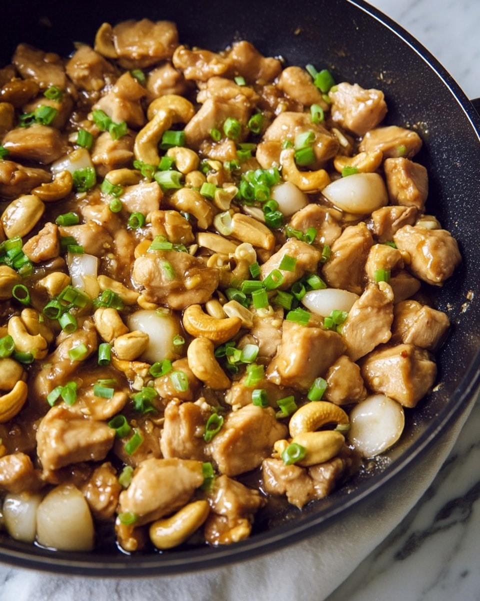 The dish is shown in a black skillet filled with small chunks of cooked chicken that are light brown and glistening, mixed with light tan cashew nuts and slices of pale white water chestnuts. Green chopped scallions are sprinkled evenly on top, adding a pop of color. The ingredients appear well-coated with a shiny sauce, and the whole dish looks moist and appetizing. The skillet is set on a white marbled surface with some part of a white cloth with blue patterns visible under it, creating a cozy setting. photo taken with an iphone --ar 4:5 --v 7