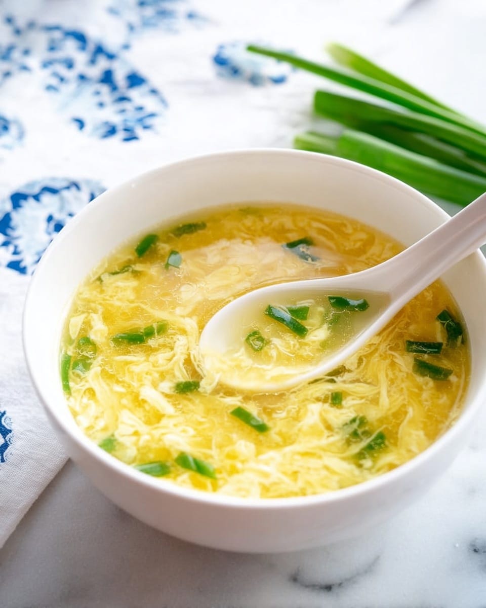 A white bowl sits on a white marbled surface filled with clear yellow broth containing soft, thin ribbons of cooked egg that create a marbled texture throughout the soup. Small pieces of bright green chopped scallions float both in the soup and on a white ceramic spoon resting in the bowl, which holds a closer view of the soup’s smooth, watery surface mixed with the fluffy egg strands. In the background, a blurred white cloth with blue patterns and fresh green scallion stalks add subtle color contrasts. Photo taken with an iphone --ar 4:5 --v 7