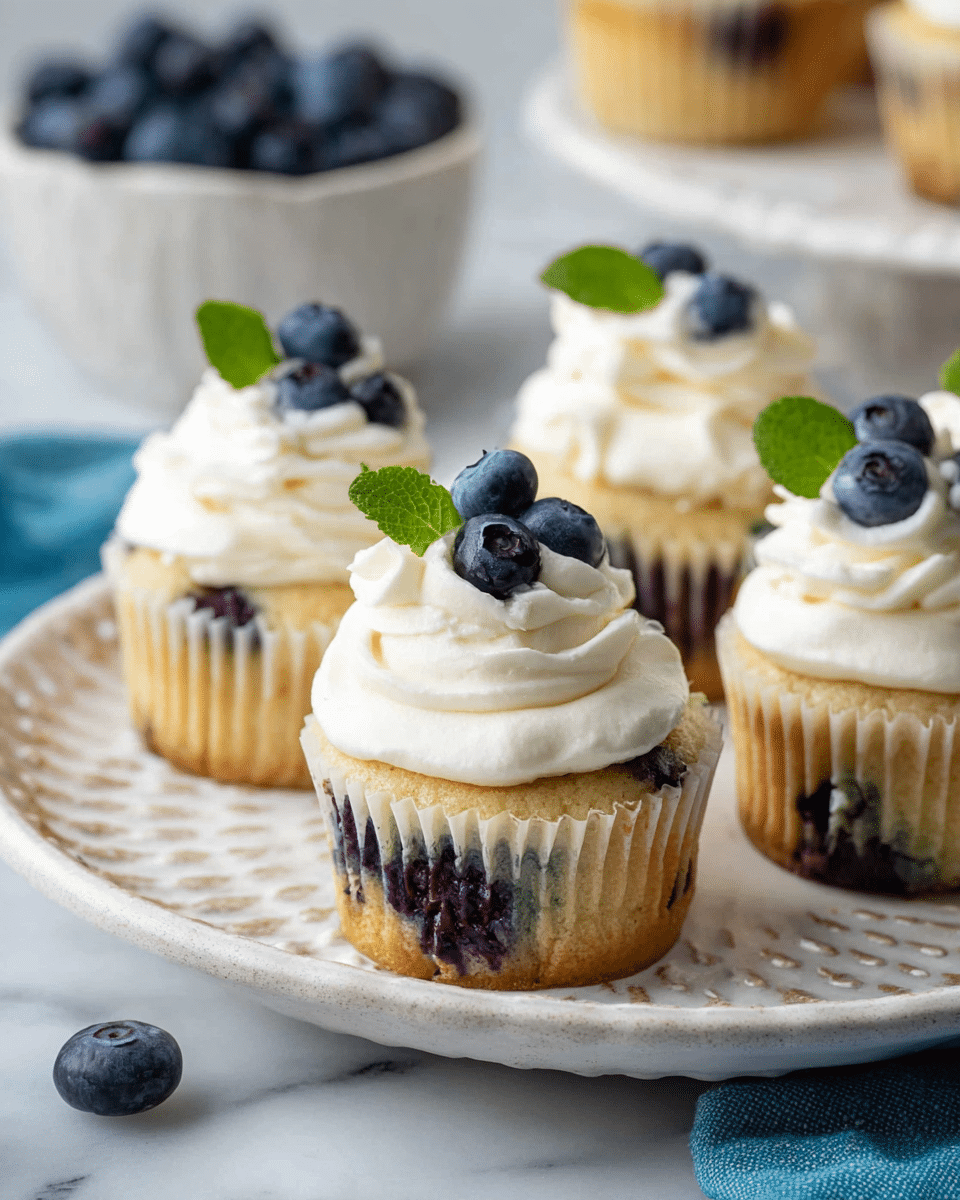 This image shows several blueberry cupcakes on a white plate with a textured edge. Each cupcake has two main layers: the base is a light golden cake with visible dark blue blueberries baked inside, and the top is a thick swirl of white cream. On the cream top, there are small dark blue fresh blueberries and a small green mint leaf placed as decoration. In the background, a white bowl filled with blueberries is partially visible, and the whole setup is on a white marbled surface with a blue cloth on one side. Photo taken with an iphone --ar 4:5 --v 7