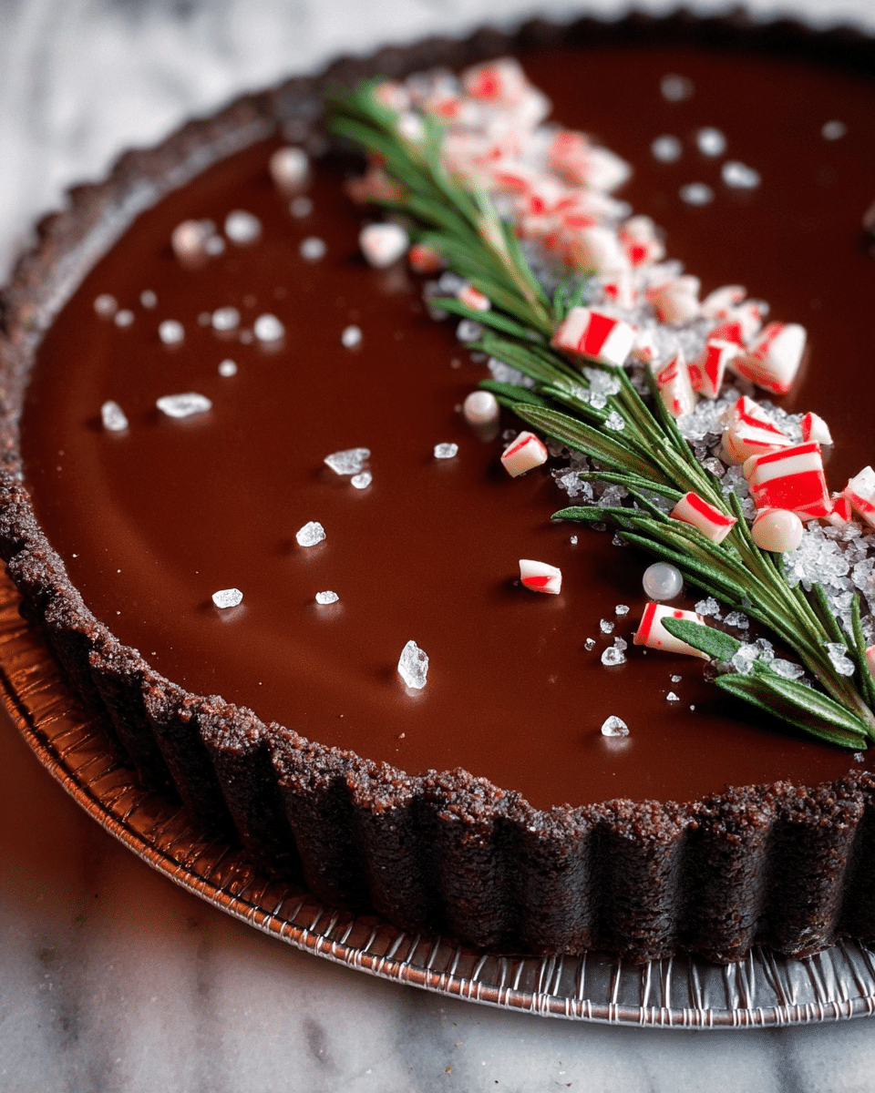 The image shows a chocolate tart with three main layers. The bottom layer is a dark brown crumbly crust that edges the entire tart. The middle layer is smooth, glossy dark chocolate filling that covers the whole tart surface. On top, there is a neat row of mint green rosemary sprigs, small white sugar beads, clear sugar crystals, and broken pieces of red and white peppermint candy spread along the tart’s center in a straight line. The tart is in a silver tart pan and sits on a white marbled texture. photo taken with an iphone --ar 4:5 --v 7