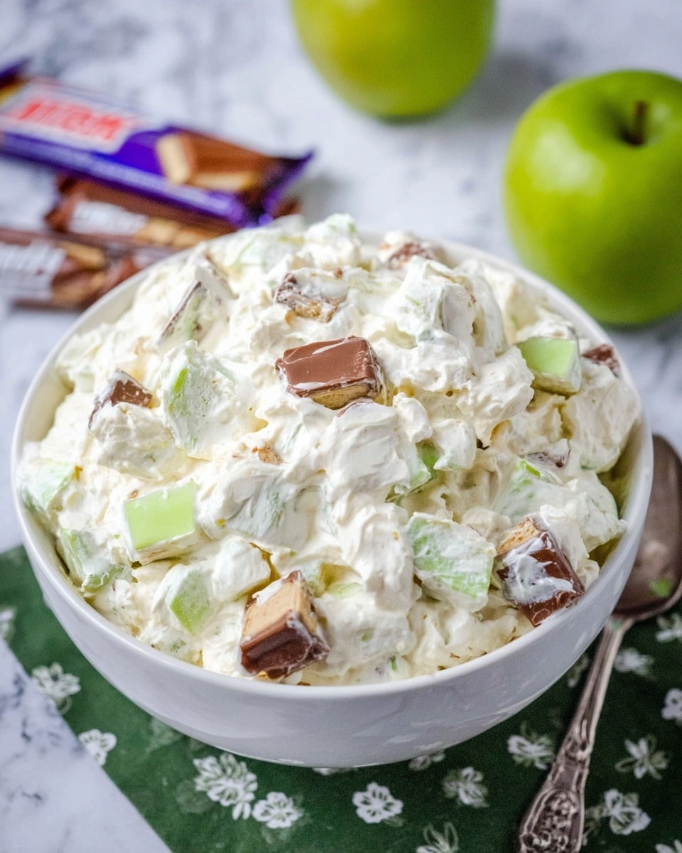 A white bowl filled with a creamy, thick white mixture with pieces of chopped Snickers candy bars and small chunks of green apple mixed evenly throughout. The mixture appears fluffy and rich, covering the candy and fruit pieces in a smooth layer. The bowl sits on a green cloth with white floral patterns, and part of a decorative silver spoon is visible next to it. The background is a white marbled surface with a whole green apple and two Snickers bars placed slightly blurred in the back. photo taken with an iphone --ar 4:5 --v 7
