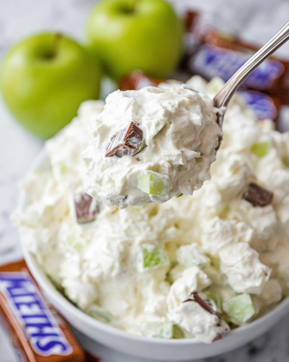 A close-up view of a spoon lifting a creamy salad scoop from a white bowl filled with a fluffy mixture that looks like whipped cream or marshmallow fluff mixed with small pieces of green apple and chunks of chocolate candy bars. The salad mixture is thick, white, and lumpy with visible soft fruit pieces and chocolate bits scattered throughout. The background is a white marbled texture with blurred green apples and wrapped candy bars visible. photo taken with an iphone --ar 4:5 --v 7