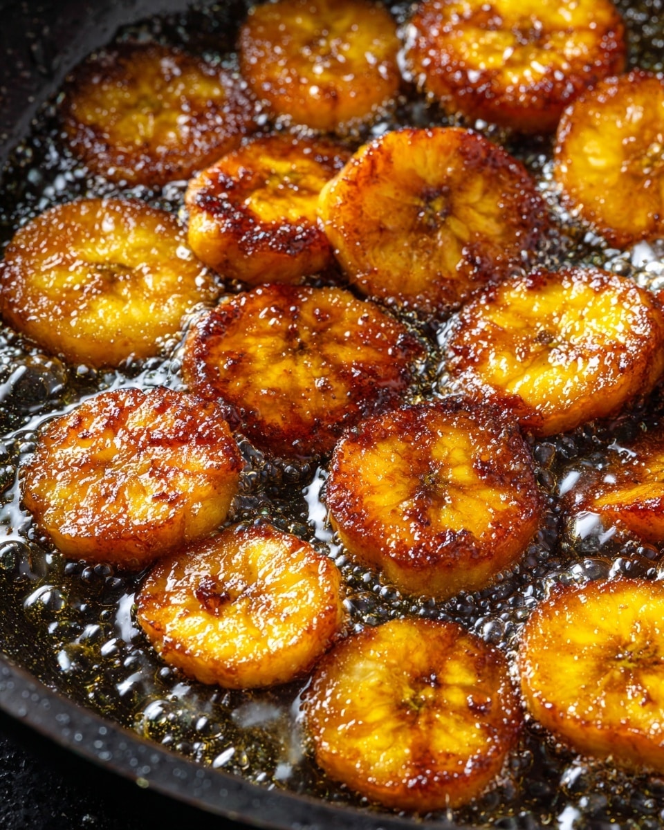 The image shows slices of plantains frying in hot oil inside a black pan. The plantain pieces are golden brown with a shiny, glazed surface from the oil, creating a crisp and caramelized texture. The slices vary slightly in size and are partially submerged in bubbling oil, which reflects light and adds a glossy finish. The close-up view focuses on the rich color and texture contrast between the crispy plantains and the smooth, shiny oil. photo taken with an iphone --ar 4:5 --v 7