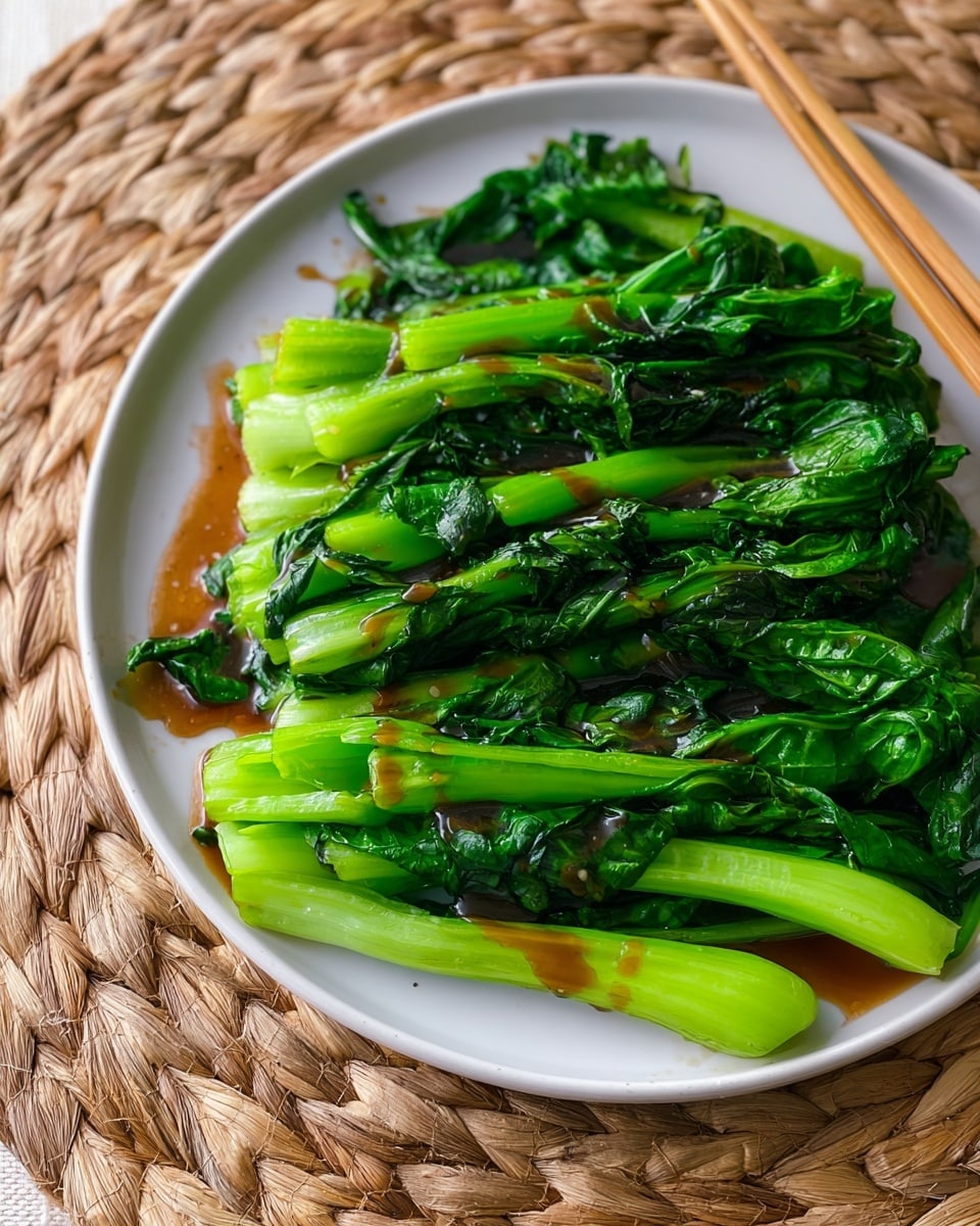 A white round plate holds a neat stack of bright green cooked leafy vegetables with thick stems arranged in two layers, with the top layer showing more stems and the bottom layer showing large leaves. The vegetables have a glossy texture with some brown sauce drizzled unevenly on top, creating a shiny contrast. The plate sits on a woven wicker-like mat, which frames the plate naturally. Beside the plate are two light wooden chopsticks. The overall image is bright with natural light, capturing fresh and healthy food. Photo taken with an iphone --ar 4:5 --v 7