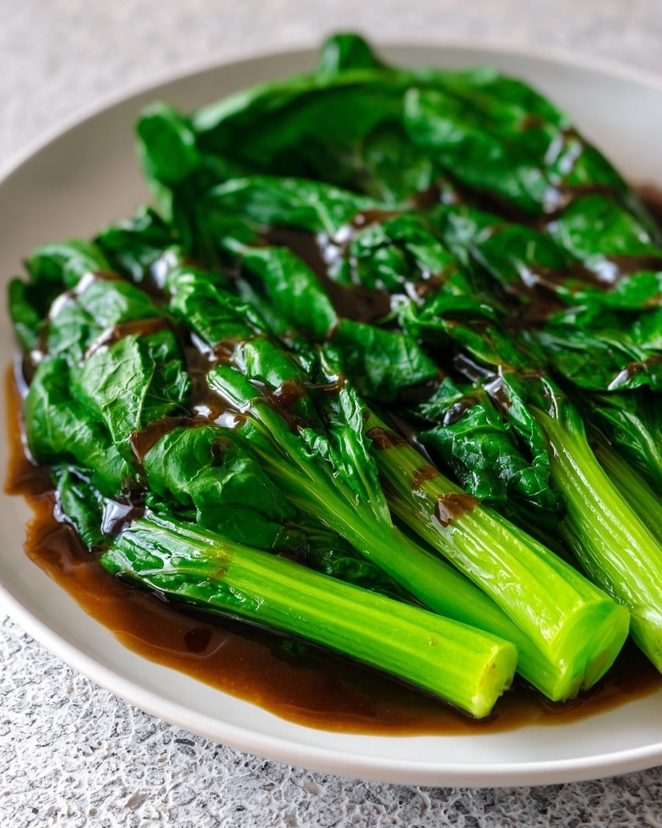 A white plate holds a single layer of bright green cooked leafy greens with thick stems, covered in a glossy dark brown sauce that pools slightly in the creases of the leaves. The leaves have a shiny, smooth texture with visible veins, and the thick green stems extend from the top side of the plate. The plate is set on a white marbled textured surface. photo taken with an iphone --ar 4:5 --v 7