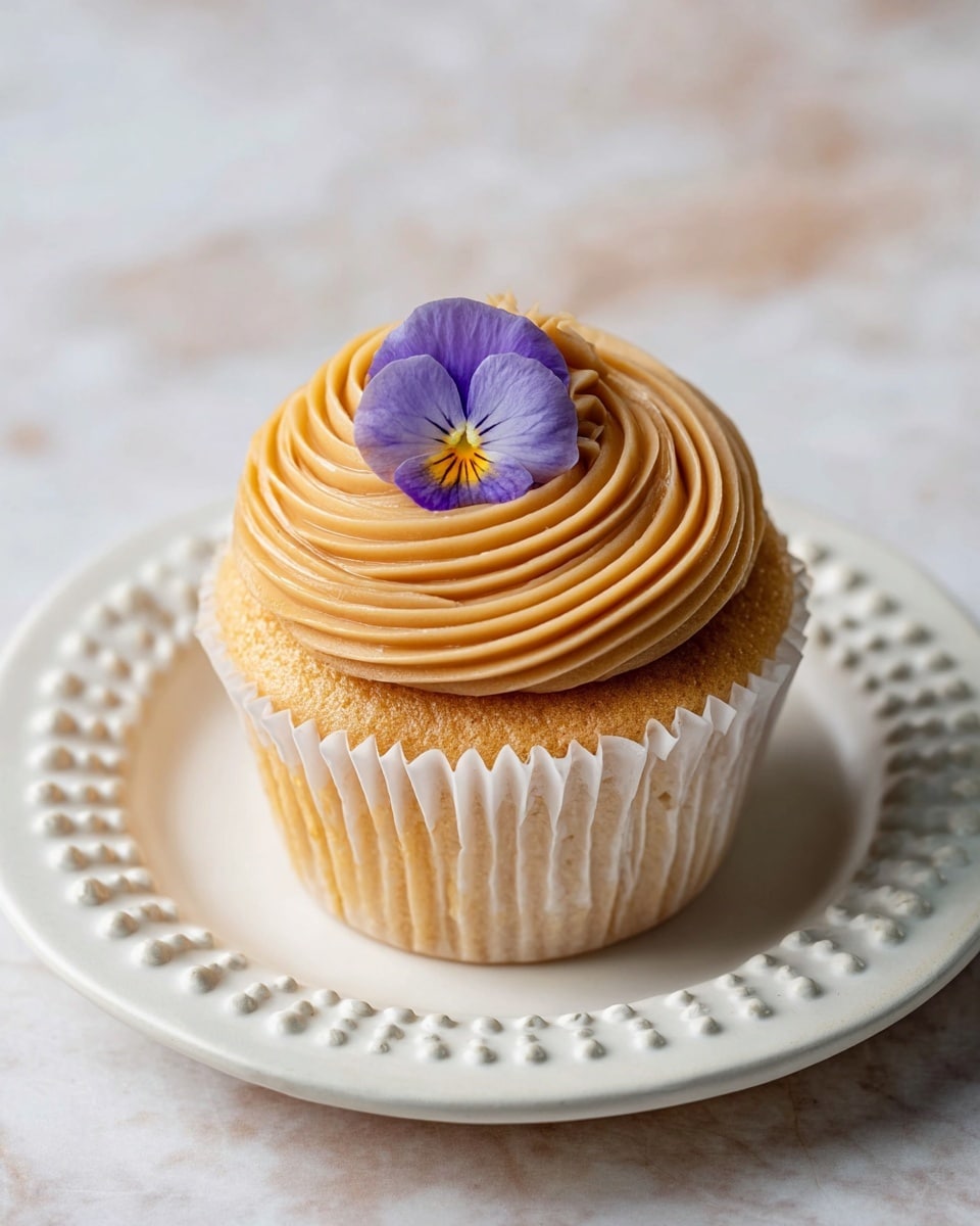 A single cupcake sits centered on a white plate with a scalloped edge and small bead-like details on the rim, placed on a white marbled surface. The cupcake has a light golden brown base with a ridged white paper liner. On top, there is a thick swirl of tan frosting, piped in tight concentric circles that cover the entire cupcake top. A delicate purple and yellow edible flower rests right at the center of the frosting swirl, adding a pop of color to the soft tan and golden tones. The overall look is simple yet elegant, with smooth textures and clean lines. photo taken with an iphone --ar 4:5 --v 7