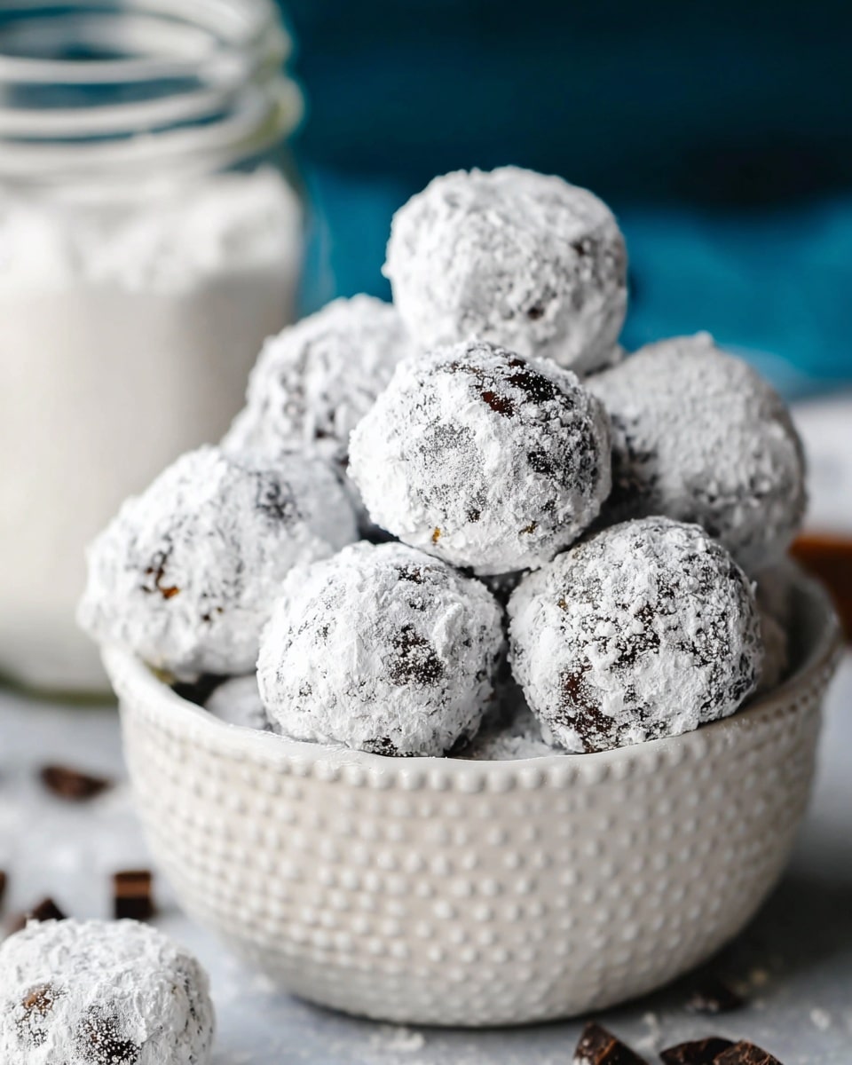 A pile of round chocolate balls covered in a thick layer of white powdered sugar fills a white bowl with a textured exterior. Each ball shows bits of dark chocolate peeking through the powdery white coating. The white marbled textured surface is scattered with small dark chocolate pieces near the bowl, while a glass jar filled with more powdered sugar is blurred in the background. The overall look is soft with a mix of powdery and rough textures. photo taken with an iphone --ar 4:5 --v 7