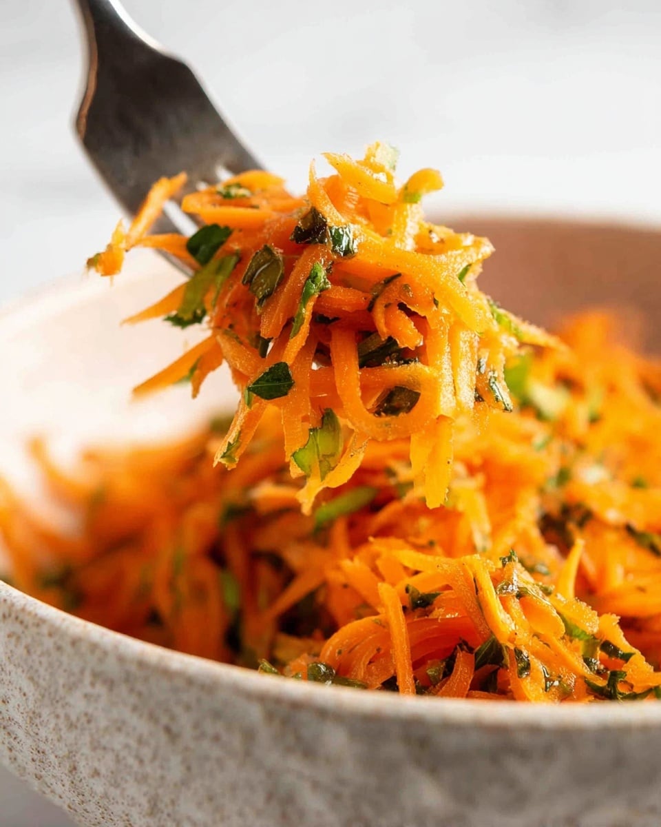 A close-up view shows a fork lifting a mix of thin orange shredded carrots and thin green herb pieces from a textured white bowl. The carrot strands are soft and glossy, layered with small dark green herb bits scattered unevenly throughout. The background is a smooth white marbled texture, which highlights the bright warm colors of the shredded carrots and herbs. Photo taken with an iphone --ar 4:5 --v 7