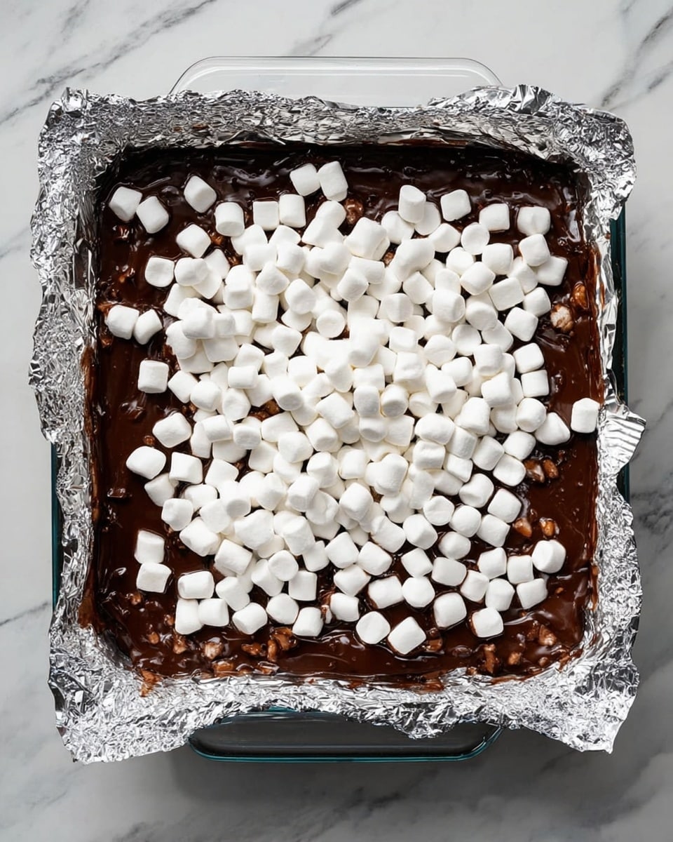 A square glass baking dish lined with crinkled aluminum foil holds a dessert before baking. The first visible layer is thick, glossy dark brown chocolate mixed with slightly visible darker chunks or nuts, covering the bottom completely. On top, an even layer of small white mini marshmallows is scattered, creating a soft, puffy texture contrast with the chocolate below. The dish sits on a white marbled surface. Photo taken with an iphone --ar 4:5 --v 7