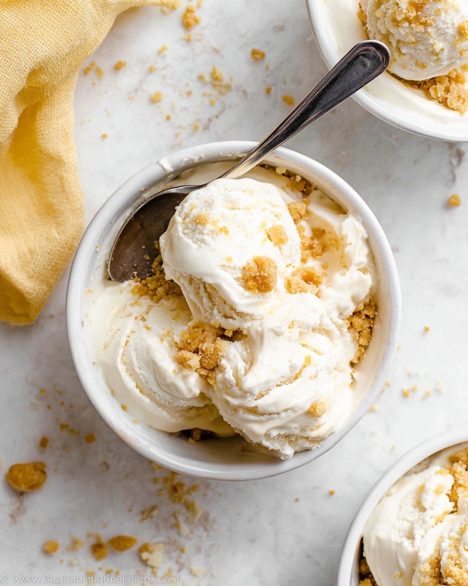 The image shows a white bowl filled with several scoops of creamy white ice cream mixed with golden crumbly pieces scattered on top and throughout. The ice cream has a soft, smooth texture with visible chunks of the crumbly layer adding roughness. A silver spoon is placed inside the bowl, resting against the side and partly buried in the ice cream. The bowl sits on a white marbled surface with crumbs scattered around it and a yellowish cloth partially visible to the left. Another similar bowl is partially seen at the top right corner. Photo taken with an iphone --ar 4:5 --v 7