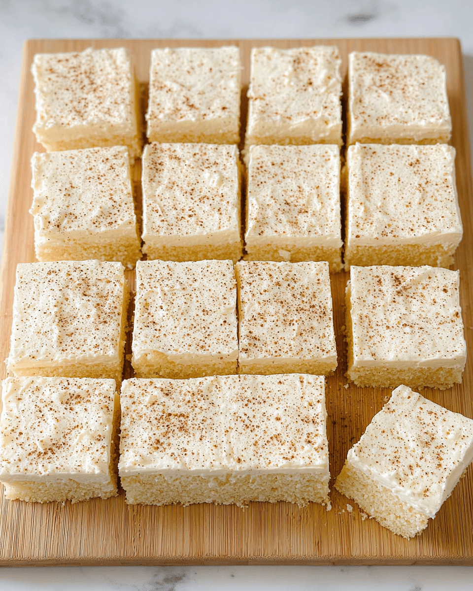 The image shows sixteen square pieces of a creamy, frosted cake arranged neatly in four rows on a light wooden cutting board, placed on a white marbled surface. Each piece has two visible layers: a pale yellow fluffy cake base and a thick, smooth white frosting layer on top. The frosting is evenly spread and sprinkled lightly with brown specks, possibly cinnamon or nutmeg, giving a slightly textured look. The edges of the cake pieces are cleanly cut, with one piece slightly pulled away from the rest on the bottom right corner. photo taken with an iphone --ar 4:5 --v 7