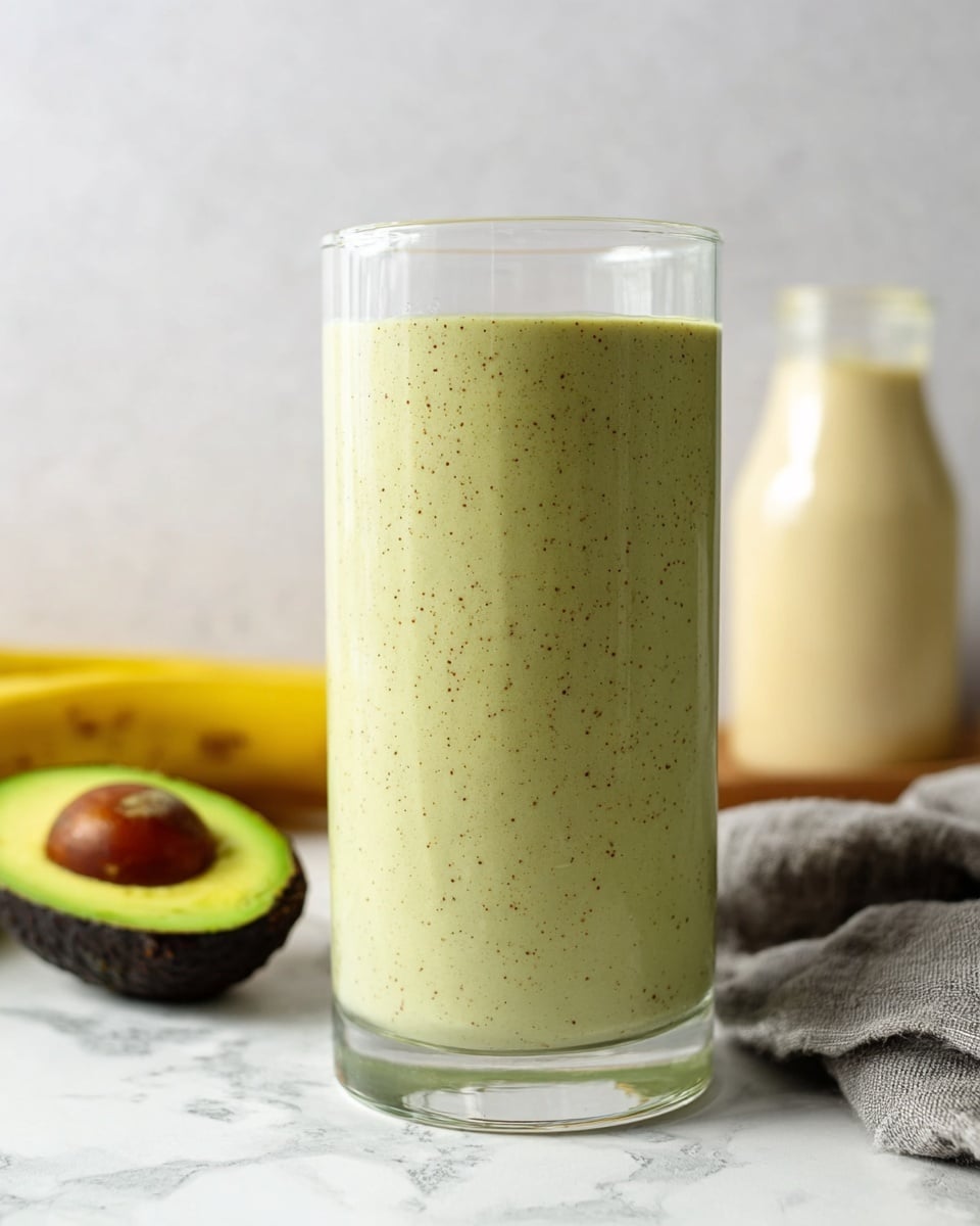A tall clear glass filled with a thick, pale green smoothie that has tiny dark specks evenly spread throughout the smooth texture, sitting on a white marbled surface; in the background, there is a halved avocado showing its bright green flesh and dark brown seed, a whole yellow banana lying horizontally, a clear bottle with light creamy liquid, and a gray cloth casually placed. Photo taken with an iphone --ar 4:5 --v 7