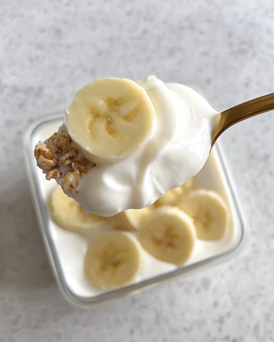 A close-up image shows a spoon holding a thick layer of white creamy yogurt with a slice of light yellow banana on top, some granola pieces peeking through the yogurt. In the background, a clear square container filled with smooth white yogurt is topped with several banana slices. The scene is set on a white marbled texture surface. photo taken with an iphone --ar 4:5 --v 7