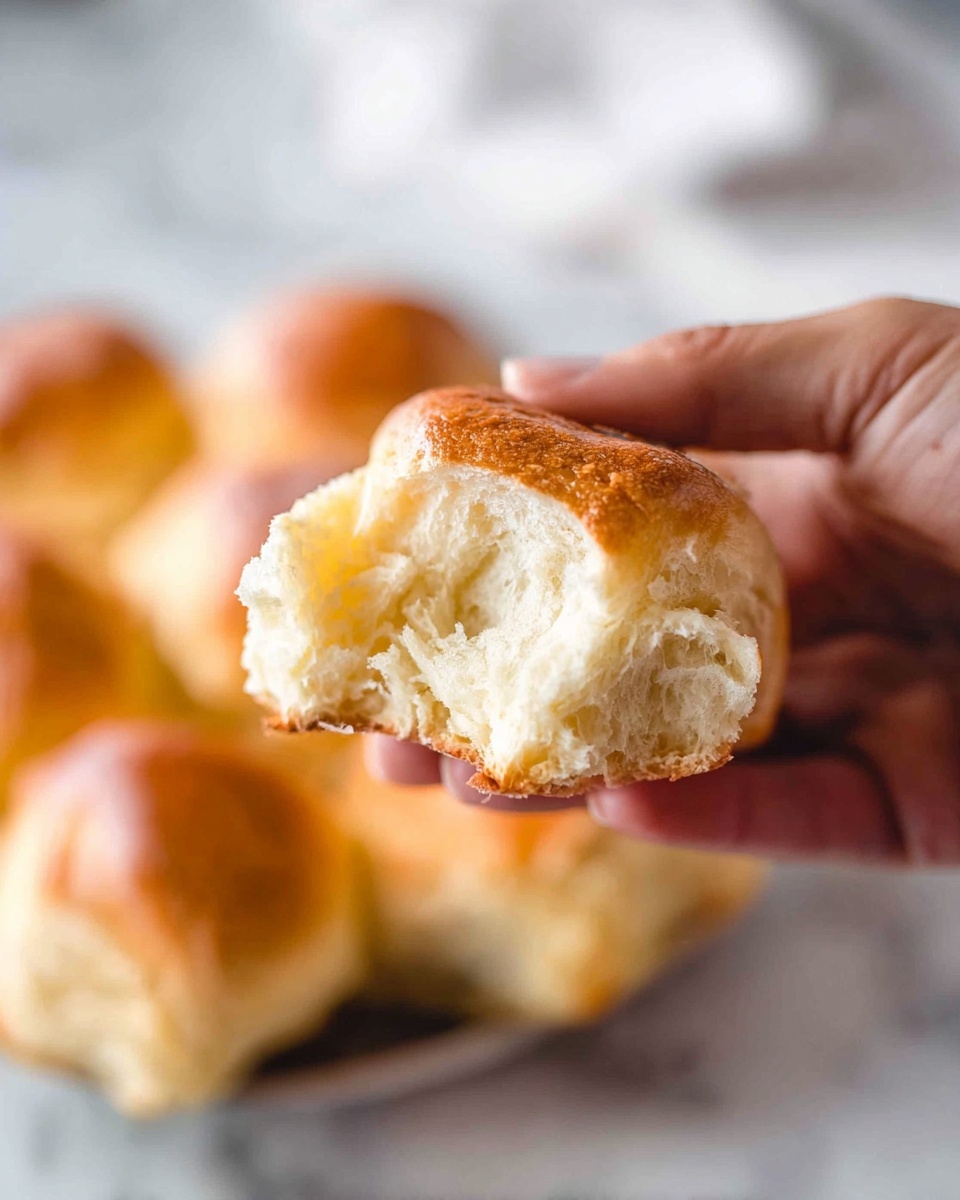 A close-up of a woman's hand holding a torn soft bread roll with a golden-brown crust and light, fluffy white inside. The torn part clearly shows the soft texture and slight gloss of the bread. In the blurred background, there are several whole bread rolls arranged together on a white marbled surface. The lighting highlights the bread's warm, inviting color and the smooth skin of the woman's hand. photo taken with an iphone --ar 4:5 --v 7