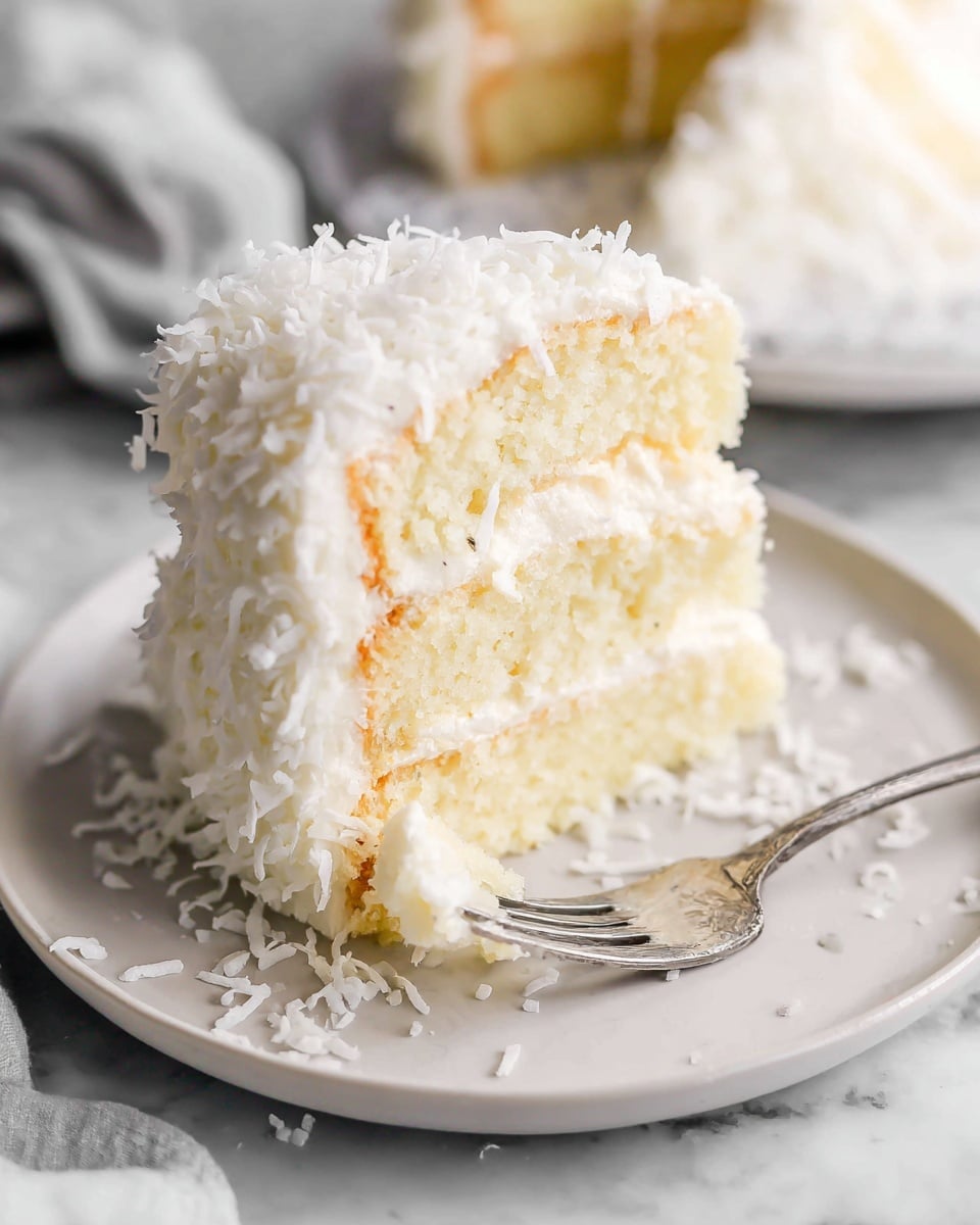 A slice of soft white layered cake sits on a white plate, with three visible layers separated by creamy, pale yellow filling. The outside is covered in fluffy white frosting and topped with shredded white coconut, which also spills onto the plate around the cake. A fork rests cutting into the front bottom layer of the cake, showing the moist texture inside. The plate is set on a white marbled surface, and the background is softly blurred to keep the focus on the cake. photo taken with an iphone --ar 4:5 --v 7