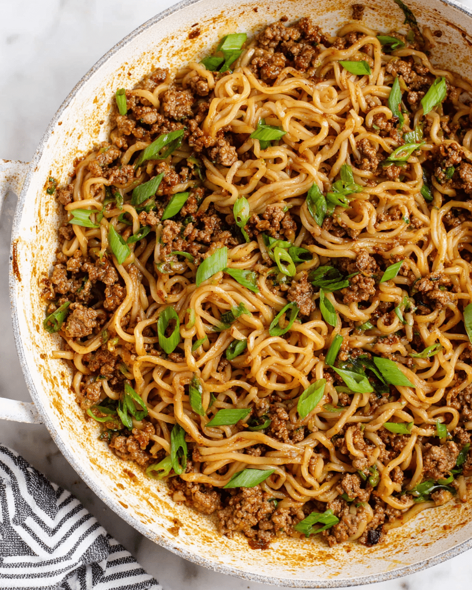 A close-up view of a white pan filled with a mix of light brown noodles and small chunks of cooked ground meat, seasoned and scattered with pieces of bright green chopped scallions. The noodles and meat are evenly mixed, showing some glistening sauce coating them. The pan has some browned marks and sauce residue around the edges, and it sits on a white marbled surface with a striped cloth partially visible on one side. photo taken with an iphone --ar 4:5 --v 7