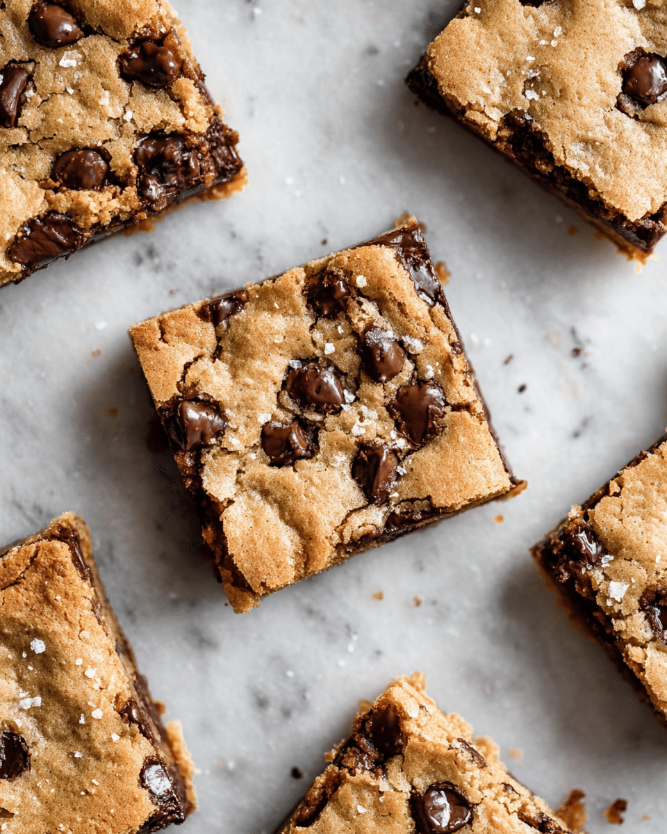 Several square cookie bars are scattered across a white marbled surface. Each bar has a golden brown top layer that looks slightly cracked and soft, dotted with large, melted, dark chocolate chips. The edges show a gooey, rich chocolate layer underneath the golden top, with some chocolate slightly oozing out. There are visible sprinkles of coarse salt on the surface of some bars, adding texture and shine. The overall look is warm, inviting, and freshly baked. photo taken with an iphone --ar 4:5 --v 7