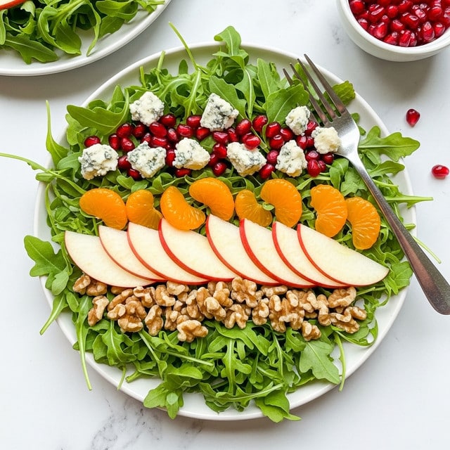 A fresh salad arranged on a white plate with three main layers: the base layer is bright green arugula leaves with some stems, the middle layer has thin slices of red apple with white inner flesh spread evenly, and scattered orange mandarin segments add a pop of color. On top, small crumbles of blue cheese with a pale blue-green color and pomegranate seeds in rich red are sprinkled along with some chopped walnuts in light brown. The salad is placed on a white marbled surface with a fork resting on the right side of the plate, and part of another white plate with more salad is visible in the background along with a small white bowl filled with red pomegranate seeds. Photo taken with an iphone --ar 4:5 --v 7
