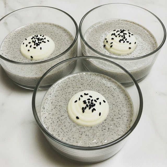 The image shows three clear glass bowls arranged close together on a white marbled surface. Each bowl contains a two-layer dessert: the bottom layer is a creamy, speckled gray mixture with a slightly grainy texture, and the top layer is a small dollop of smooth white cream placed in the center. The cream on each bowl is decorated with tiny black seeds scattered on top, adding contrast. The overall look is simple and clean with soft colors and clear glass that reveals the layers inside. photo taken with an iphone --ar 4:5 --v 7