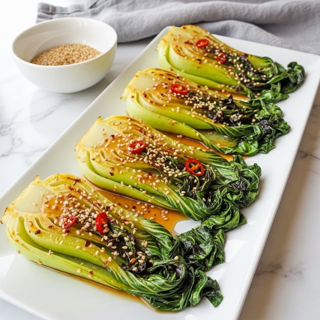 The image shows four cooked baby bok choy arranged in a row on a long, white rectangular plate. Each piece has a bright yellow-green top layer with a glossy, slightly charred texture, and dark green, wilted leaves at the bottom. The bok choy is covered in a shiny brown sauce with visible small sesame seeds and bits of red chili flakes sprinkled evenly over the top. The plate sits on a white marbled surface, and a white bowl with light brown sesame seeds is placed nearby. A soft gray cloth is in the background. photo taken with an iphone --ar 4:5 --v 7