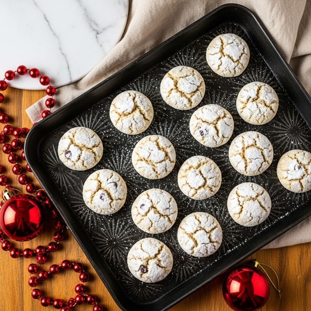The image shows a dark metal baking tray with a textured starburst pattern, holding twelve round cookies dusted lightly with white powdered sugar. Each cookie is light tan with small darker spots and a slightly craggy surface, giving a soft, crumbly look. The tray is placed on a wooden table with a string of large red beads and a red shiny ornament nearby. In the background, there is a white marbled texture visible under a beige cloth, adding a soft contrast to the warm and festive setting. Photo taken with an iphone --ar 4:5 --v 7