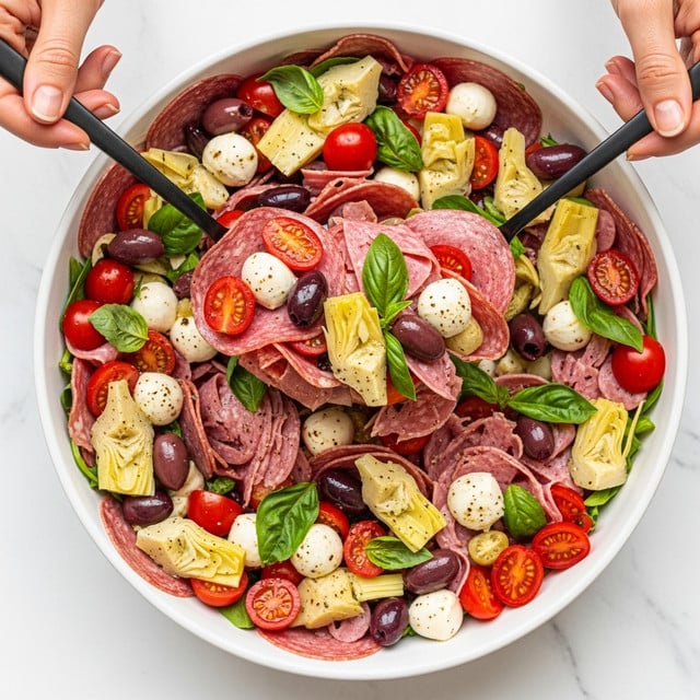 A large white bowl filled with a colorful Italian salad showing many thinly sliced layers of pink salami and prosciutto mixed with small white mozzarella balls, bright red halved cherry tomatoes, yellow-green marinated artichoke pieces, dark purple olives, and scattered green basil leaves. Two black spoons are lifting a mix of the layers, and woman’s hands hold the spoons against a white marbled background. The salad looks fresh and vibrant with a mix of soft, firm, and leafy textures. photo taken with an iphone --ar 4:5 --v 7