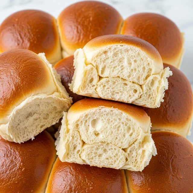 The image shows a close-up of many soft, golden brown rolls stacked together. Each roll has a smooth, lightly browned crust with a soft, slightly airy texture inside. Some rolls display their inner layers, revealing a light, fluffy, creamy-yellow dough with small air pockets, folded softly in two to three layers. The rolls have a slightly shiny, baked surface with an inviting warm color, showing slight crisp edges on some sides. The background is a simple white marbled texture. Photo taken with an iphone --ar 4:5 --v 7