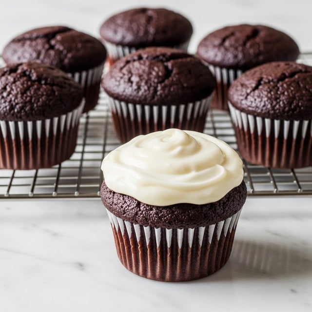 This image shows a group of seven chocolate cupcakes placed on a metal cooling rack over a white marbled surface. Six of the cupcakes have a dark brown, slightly cracked top, and are wrapped in white paper liners. One cupcake stands out in the foreground with a smooth, thick, and creamy white frosting layer evenly spread on top, also in a white paper liner. The texture of the frosting is glossy and soft, covering the entire top of the cupcake without dripping. The rest of the cupcakes have a crumbly surface with no frosting. Photo taken with an iphone --ar 4:5 --v 7