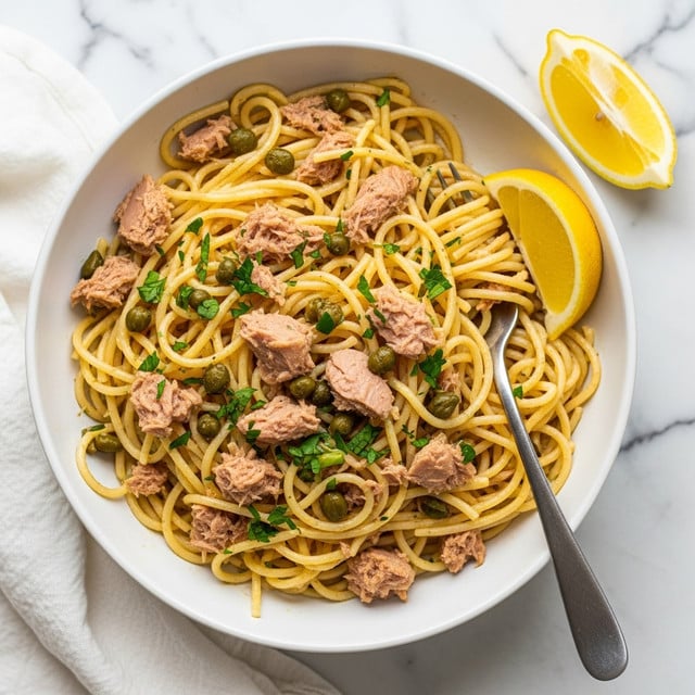 A bowl of spaghetti pasta mixed with small chunks of tuna, scattered green capers, and sprinkled chopped parsley on top. The pasta is a light yellow color, coated in olive oil, with pieces of tuna in light brown, and green capers adding texture and color contrast. A silver fork is placed inside the bowl on the right side, and two lemon wedges rest beside the pasta on the right edge. The bowl is white and set on a white marbled surface with a white cloth beneath it. photo taken with an iphone --ar 4:5 --v 7