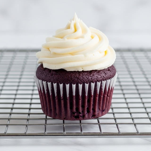 A single cupcake is shown close up, sitting on a metal cooling rack over a white marbled texture. The cupcake has one layer of dark purple cake with a moist texture and small air holes. It is wrapped in a white paper liner that has ridged edges. On top, there is one thick layer of white frosting piped in a smooth swirl with soft peaks, centered neatly on the cake. The lighting highlights the texture of the cake and the creamy frosting. photo taken with an iphone --ar 4:5 --v 7