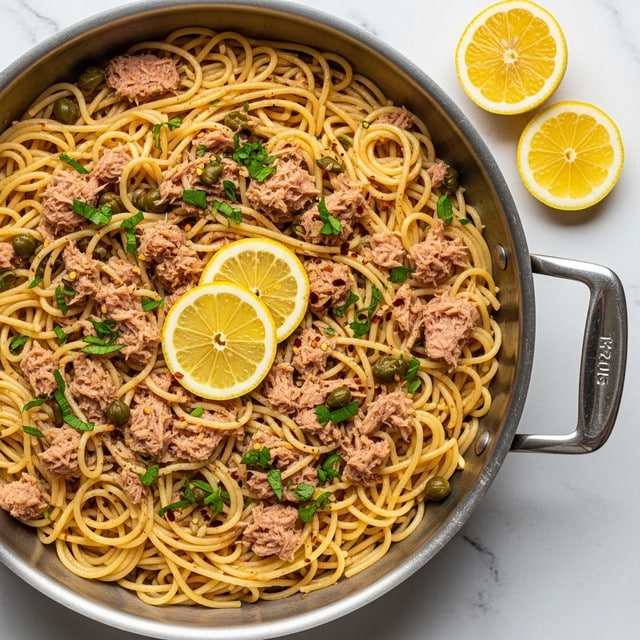 A large silver pan filled with a dish of spaghetti pasta lightly coated in oil, mixed with small chunks of pink tuna, dark green capers, and sprinkled with chopped green herbs and red pepper flakes. Two thin slices of lemon rest on top of the noodles near the center. The pan sits on a white marbled textured surface, with two lemon slices placed off to the side nearby. The image shows a close-up view emphasizing the textures and colors of the pasta and ingredients, photo taken with an iphone --ar 4:5 --v 7