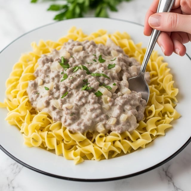 A white plate with a black rim holds a dish of wide yellow egg noodles spread out as the base layer, soft and curly in texture. On top is a thick layer of creamy, pale beige ground meat sauce with visible small chunks of browned meat and bits of onion, coated in a smooth white sauce. Sprinkled green herbs are scattered across the top, adding color. A woman's hand is holding a silver spoon, stirring into the noodles and sauce from the right side of the image. The background is a white marbled texture with some blurred green herbs behind the plate. Photo taken with an iphone --ar 4:5 --v 7
