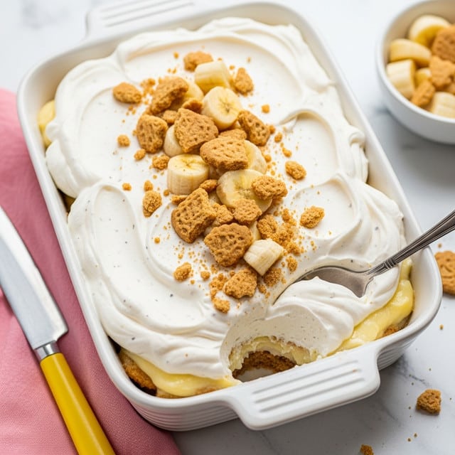 A white ceramic rectangular dish filled with a layered dessert visible on the edges, topped with a thick and fluffy layer of whipped cream that is smooth and white with small specks of vanilla. Scattered over the whipped cream are small chunks of banana and crumbled light brown cookie pieces for texture. A silver spoon is partially dipped into the right side of the whipped topping, lifting some of the dessert underneath. The dish is placed on a white marbled surface, with a pink napkin and a yellow knife beside it, and a small white bowl with more banana and cookie pieces partly visible nearby. Photo taken with an iphone --ar 4:5 --v 7