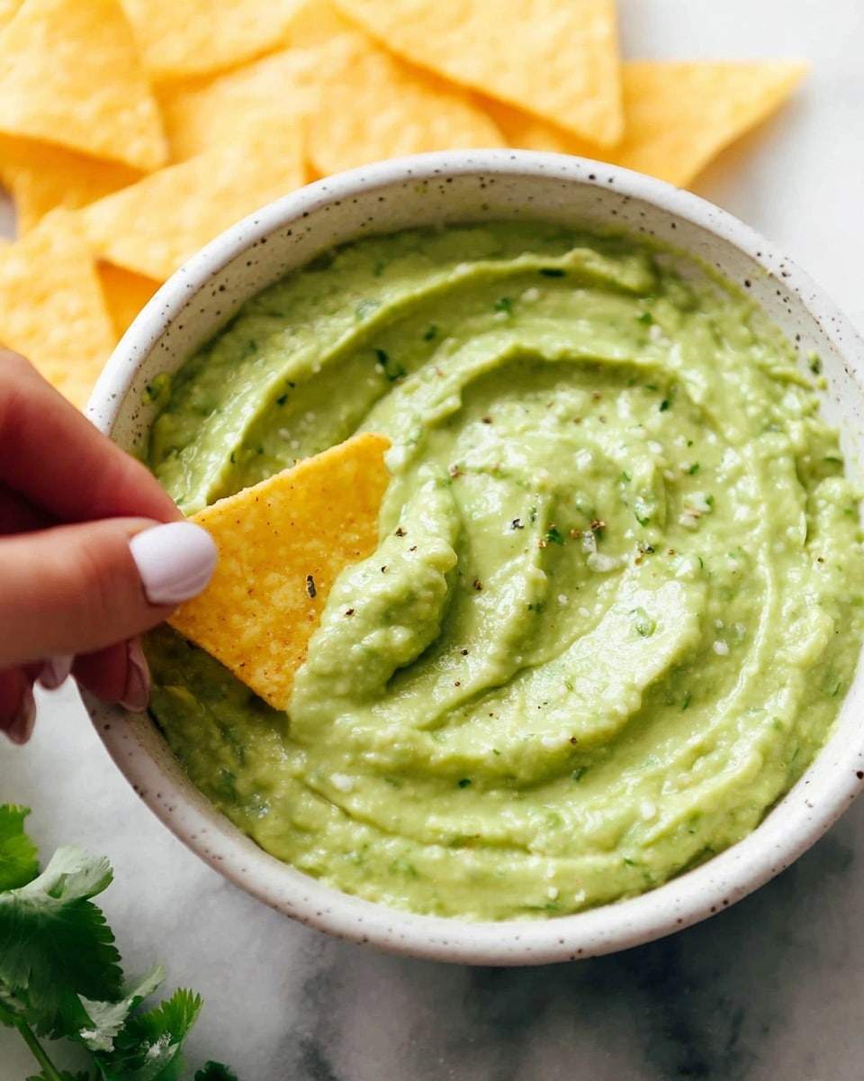 A close-up image of a creamy green guacamole dip with a smooth, slightly chunky texture in a white speckled bowl, showing gentle swirls and small pieces of herbs or spices mixed in. A yellow taco chip is dipped halfway into the guacamole, held by a woman's hand with neatly manicured light pink nails. The background features a white marbled surface, and there are triangular yellow taco chips placed slightly out of focus behind the bowl. Some green herb leaves peek in from the bottom left corner. Photo taken with an iphone --ar 4:5 --v 7