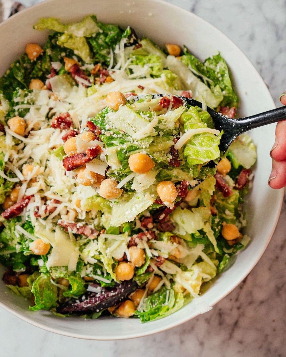 The image shows a close-up of a mixed salad in a white bowl. The salad has several layers starting with dark and light green leafy lettuce mixed throughout. There are small orange chickpeas scattered evenly across the salad. Thin strips of reddish meat or sausage add contrast, mixed with shredded white cheese. The textures range from crunchy lettuce to soft cheese and chickpeas. A black spoon lifts part of the salad, held by a woman's hand on the right edge of the bowl. The background surface is a white marbled texture. Photo taken with an iphone --ar 4:5 --v 7