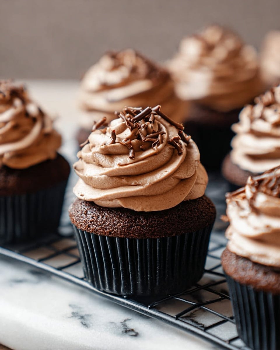 A close-up view of multiple chocolate cupcakes placed on a white marbled surface with a black cooling rack. Each cupcake has two main layers: the bottom layer is a dark brown, moist chocolate cake in a black cupcake liner, and the top layer is a soft, swirled mound of creamy, light brown chocolate frosting. The frosting is decorated with small, thin, curled chocolate shavings scattered on top. The focus is mainly on one cupcake in the center, with others softly blurred in the background. The lighting is natural, highlighting the rich textures and smoothness of the frosting. Photo taken with an iphone --ar 4:5 --v 7