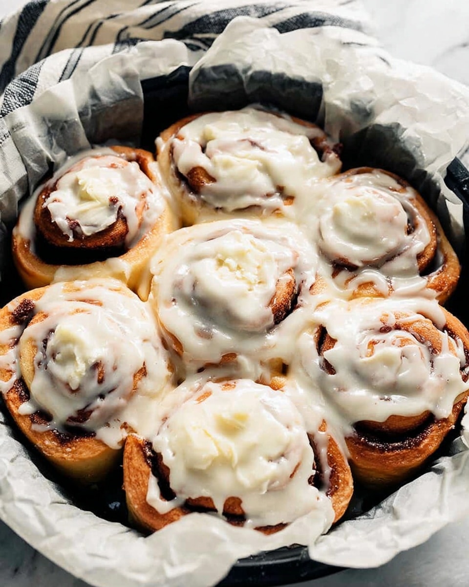 A black round pan lined with crumpled white parchment paper holds seven cinnamon rolls closely stacked together. Each cinnamon roll has a light brown dough base with dark brown cinnamon swirls visible beneath a thick, creamy white icing that covers most of each roll unevenly, showing some texture and slight melting. The pan rests on a white marbled surface with a soft fabric napkin with black stripes visible in the background. photo taken with an iphone --ar 4:5 --v 7