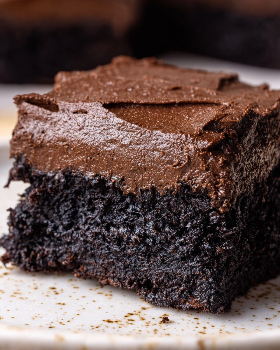 A close-up view of a chocolate dessert showing two thick layers: the bottom layer is dense, dark black-brown cake with a rough, moist texture, and the top layer is a thick, smooth, and creamy dark chocolate frosting with a slightly shiny surface and a few small air holes. The dessert is on a white plate with small brown spots, placed on a white marbled texture. Photo taken with an iphone --ar 4:5 --v 7