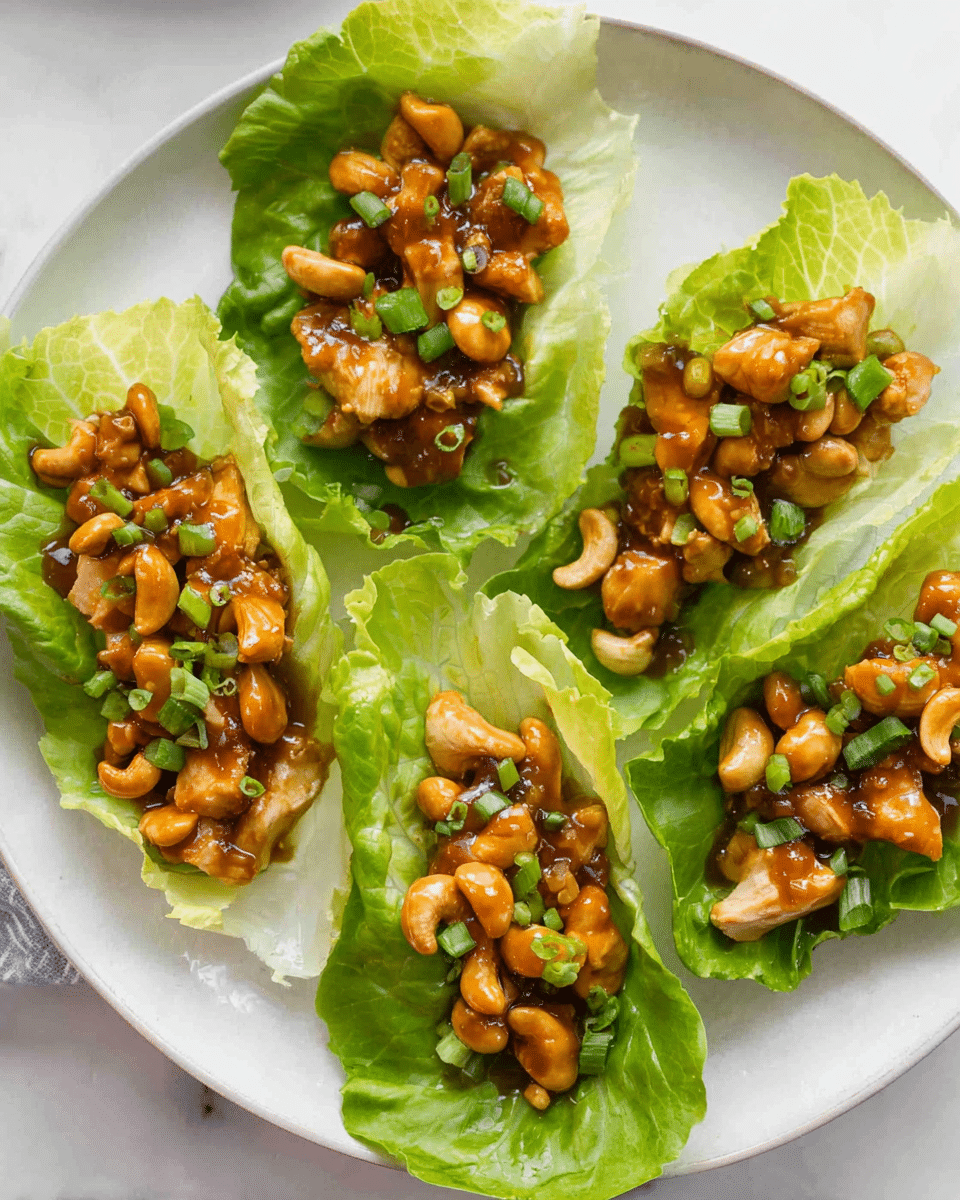 A white plate holds five lettuce cups filled with a glossy mix of chicken pieces and whole cashews coated in a dark brown sauce. Each cup shows a bright green leaf of lettuce with a crinkled texture, forming a base for the chunky chicken and nuts. The chicken pieces are light brown and shiny, while the cashews add a smooth light tan contrast. Small slices of green onions are sprinkled on top, adding a fresh green color to the dish. The plate sits on a white marbled surface. photo taken with an iphone --ar 4:5 --v 7