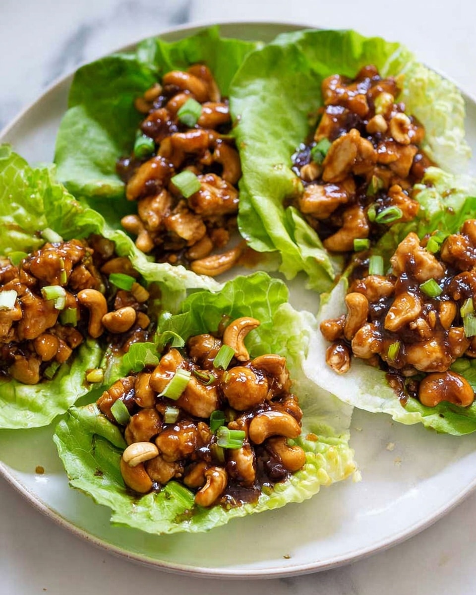 Five small green lettuce leaves are placed on a white plate with a white marbled surface underneath. Each lettuce leaf holds a mix of small cooked chicken pieces and whole cashew nuts coated in a shiny brown sauce. The mixture fills the center of each leaf, with some fresh green chopped scallions sprinkled on top for color. The textures include the crisp lettuce, the smooth sauce, the tender chicken, and the crunchy cashews mingled together. photo taken with an iphone --ar 4:5 --v 7