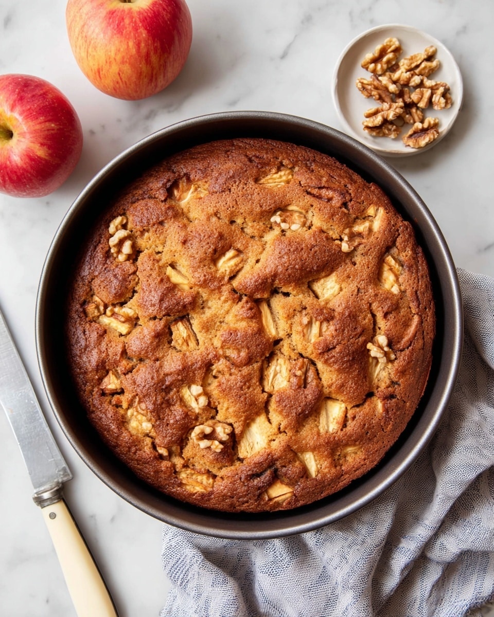 A round, golden-brown apple cake in a dark round pan, with a slightly bumpy top full of small apple chunks and scattered walnut pieces, giving a rough textured look. The cake's surface is warm brown, showing some darker spots where it is baked crispier. Next to the pan, there is a small white plate with larger walnut pieces and two whole red apples placed below the pan. A knife with a cream-colored handle lies on the white marbled surface next to the pan, along with a gray cloth with thin blue stripes partially under the pan. photo taken with an iphone --ar 4:5 --v 7